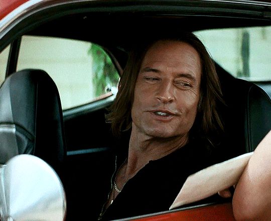 The actor Josh Holloway in the tv show “Duster”, sitting in a car and smiling up at someone who swats his shoulder with an envelope.