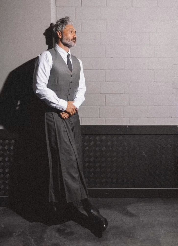 Taika Waititi wearing a gray skirt, vest and tie, leaning against a white brick wall in a hallway (2022)
