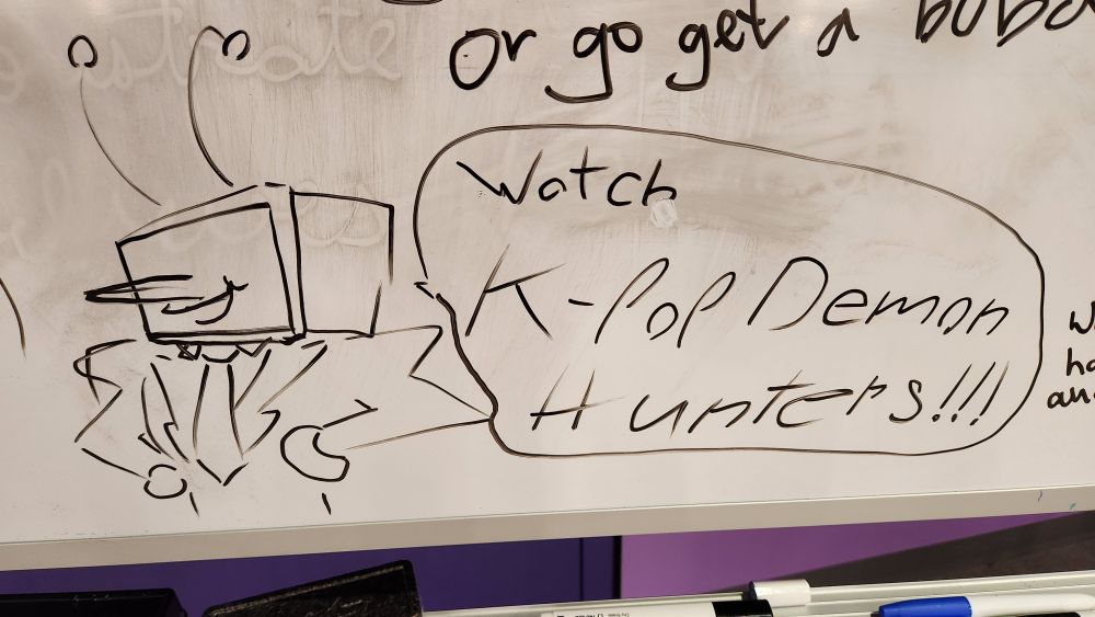 A doodle of Tenna from Deltarune in black marker on a whiteboard. A speech bubble beside Tenna says "Watch K-pop Demon Hunters!!!"