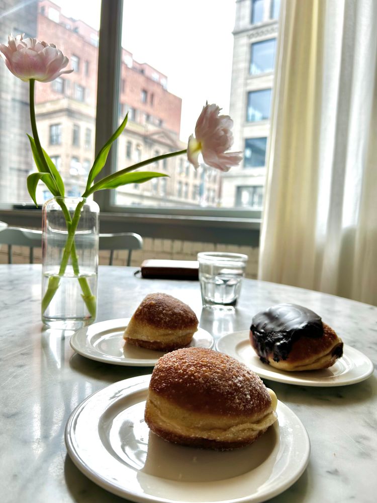 Three doughnuts, each on a plate. All are sitting on a white marble table with two pink tulips behind them and a big window view out onto the street behind it all. 