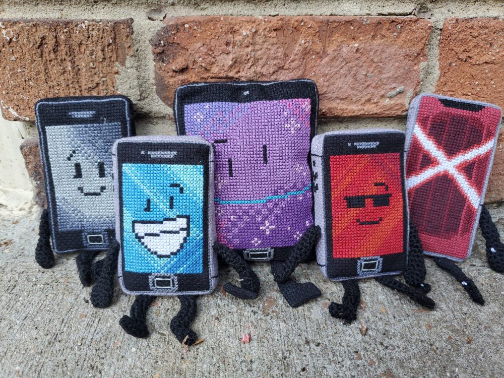 Five handmade plushies of MePhones 3GS, 4, 4S, X, and MePad are propped up against the wall. They're cross stitched, except for their crocheted limbs. 