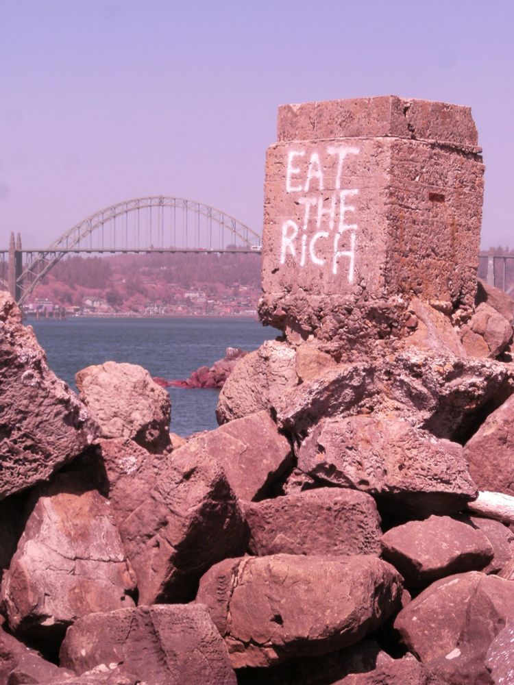 A large concrete block atop a pile of rocks, (part of Newport, Oregon's South Jetty) has the words "Eat the Rich" spray painted in white block letters.  The Yaquina Bay bridge spans the water in the background.  The camera was modified to capture more infrared light, giving the photo an otherworldly pink hue, especially the algae on the rocks and the hills back behind the bay.