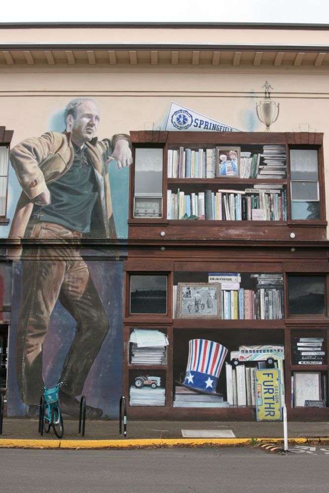 A two story Mural of author Ken Kesey leaning against his bookshelf, which is stuffed with books, photos and keepsakes, including a large Uncle Sam style top hat, Oregon license plate, from his bus Furthur, and a miniature version of the bus done in the signature Merry Prankster marbled dip-paint style. A trophy and a Springfield pennant sit atop.  Some of the bookcase is also composed of the windows of the building the mural is on.  On the sidewalk, a single light blue cruiser bicycle is parked at a bike rack at Kesey's feet.