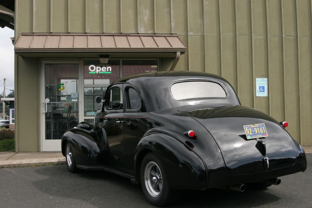 Another angle, of the back of the car reflecting the cloudy sky in its polished black paint. The corpo sandwich shop is Open, per its lit up sign in the window, its drab vertical metal siding provides contrast to the soft curves of the classic car.