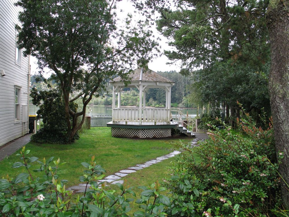 A path of stones leads to a small white gazebo which sits near a river. The whole scene is framed in foliage 
