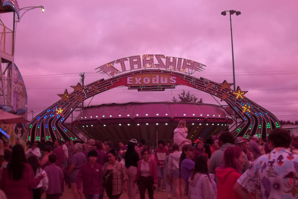  The crowd at a carnival in front of a flying saucer shaped ride under its sign reading "Starship Exodus"  There is a pinkish hue due to the camera being modified to capture infrared light.