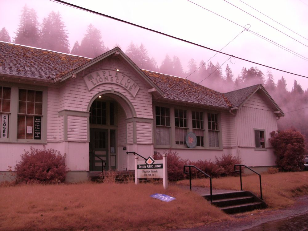 Full spectrum infrared photo of Siuslaw Public Library, Mapleton Branch.  An older sign above the door says Mapleton Lions Club.  Trees above the building's slightly mossy roof are shrouded in mist.   