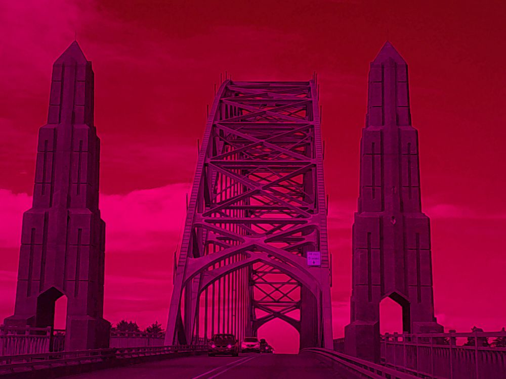 Cars pass under a bridge with a concrete column towering on each side, viewed front-on from the other side of the road.  The sky is tinted red with pink clouds, while the concrete pillars and steel bridge works have a  purple hue.