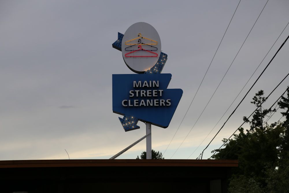 A blue sign, oddly shaped, reads "MAIN STREET CLEANERS". Above there are three neon coat hangers: yellow, blue and red in a white oval, an arrow lit with white dots bisects and connects the two sections of the sign.