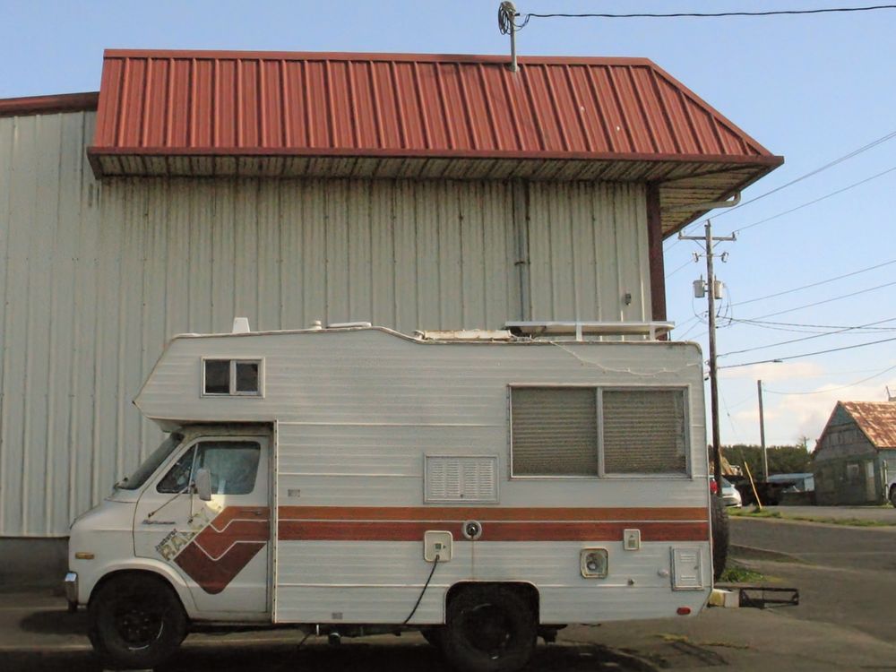 An older, smallish RV, mainly white with brown stripes running along the side sits in front of a wall with horizontal metal siding, white with a brown roof.  