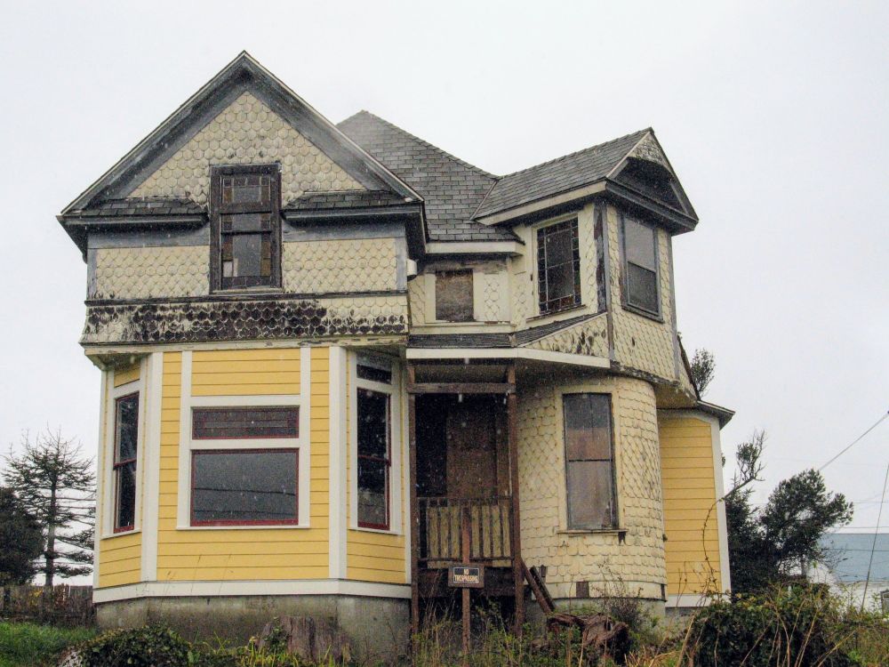 An historic house sits mid-renovation in the rain.  Parts of the exterior have been replaced with modern yellow siding, presumably to match the original color of the faded yellow shingles that still cover most of the structure.