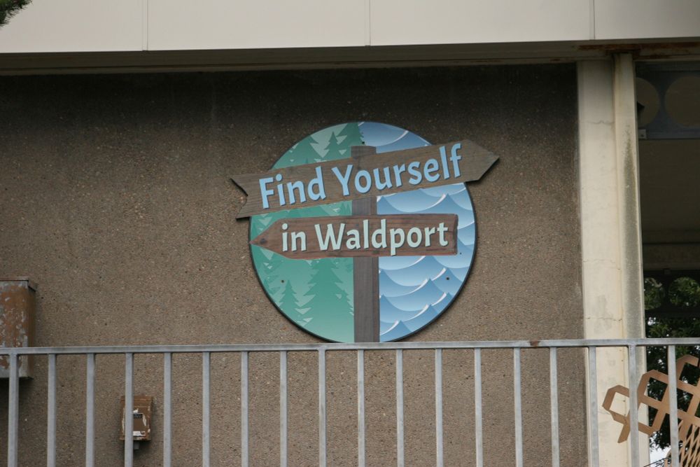A sign across the parking lot that reads "Find Yourself in Waldport". Flat and mostly round, but depicting an old signpost, "find Yourself on top, pointing right and "in Waldport" below, pointing left, with waves on one side and trees on the other, referencing Waldport's other slogan: "where the forest meets the trees"