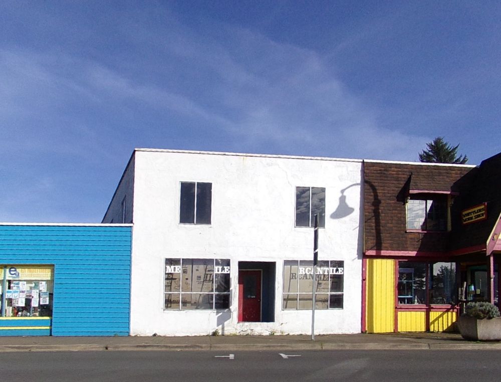 A vacant storefront in a boxy white building with a bright blue wall on its left and a red and yellow storefront with a faux shingled rooftop on the right.  "MERCANTILE" was on both lower windows, but letters are now missing where panes were replaced, a recessed red door in between them has no steps to reach it.  Wispy clouds are above and reflected in the upper windows.  An unseen lamp casts a shadow.  