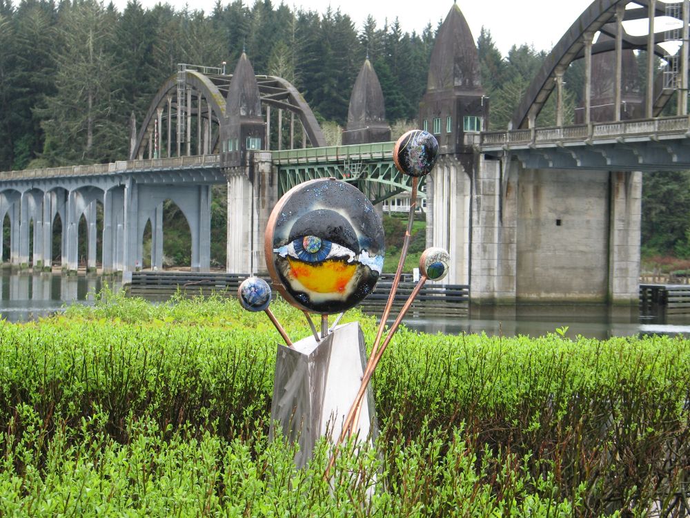 Psychedelic space eyeballs sculpture rises from amidst shrubbery with the Siuslaw River Bridge in the background.  The muted colors of the bridge and trees behind it contrast with the brighter colors of the foliage and sculpture in the foreground.