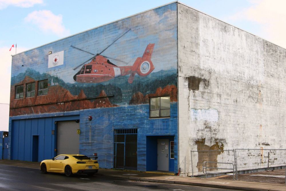 A large boxy building housing the Seawater Seafood Co., noted by a small sign on the front of the building which is otherwise completely covered with a mural of a Coast Guard helicopter over water with hills and clouds in the background, which are similar to the real clouds above the building.  The right third of the frame is taken up by the blank side wall, showing some wear and tear.  A yellow sports car is the lone vehicle parked on the street in front.  