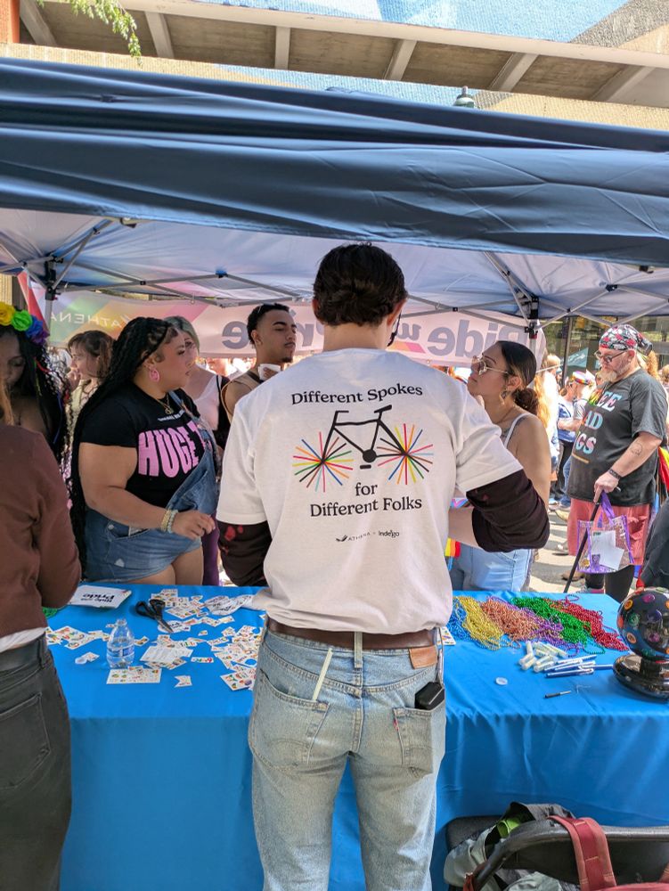 Someone working at the ride for Pride booth, rear view. The back of their shirt says "different spokes for different folks" and has a design of a bike with rainbow wheel spokes