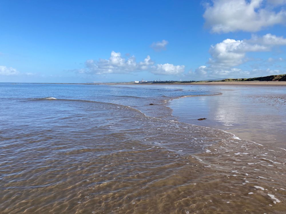 A wide beach shot with low waves trickling across the sand towards dunes and distant buildings