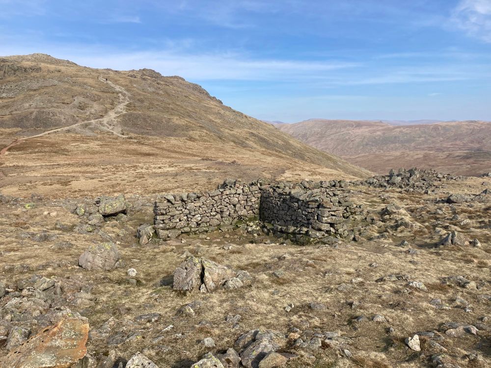 A drystone shelter cross (literally 4 drystone walls in the shape of a cross creating sheltered rest areas for walkers in the four quarters) is nestled near the bottom of Esk Hause, a dip between mountains.