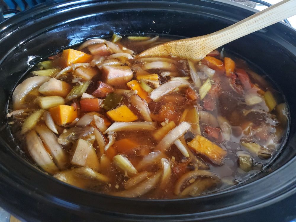 Photograph of hearty vegetable soup in a slow cooker with a wooden spoon poised to stir the soup