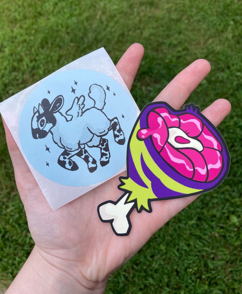 hand holding two stickers, a goat and green and purple meat. grass in the background 