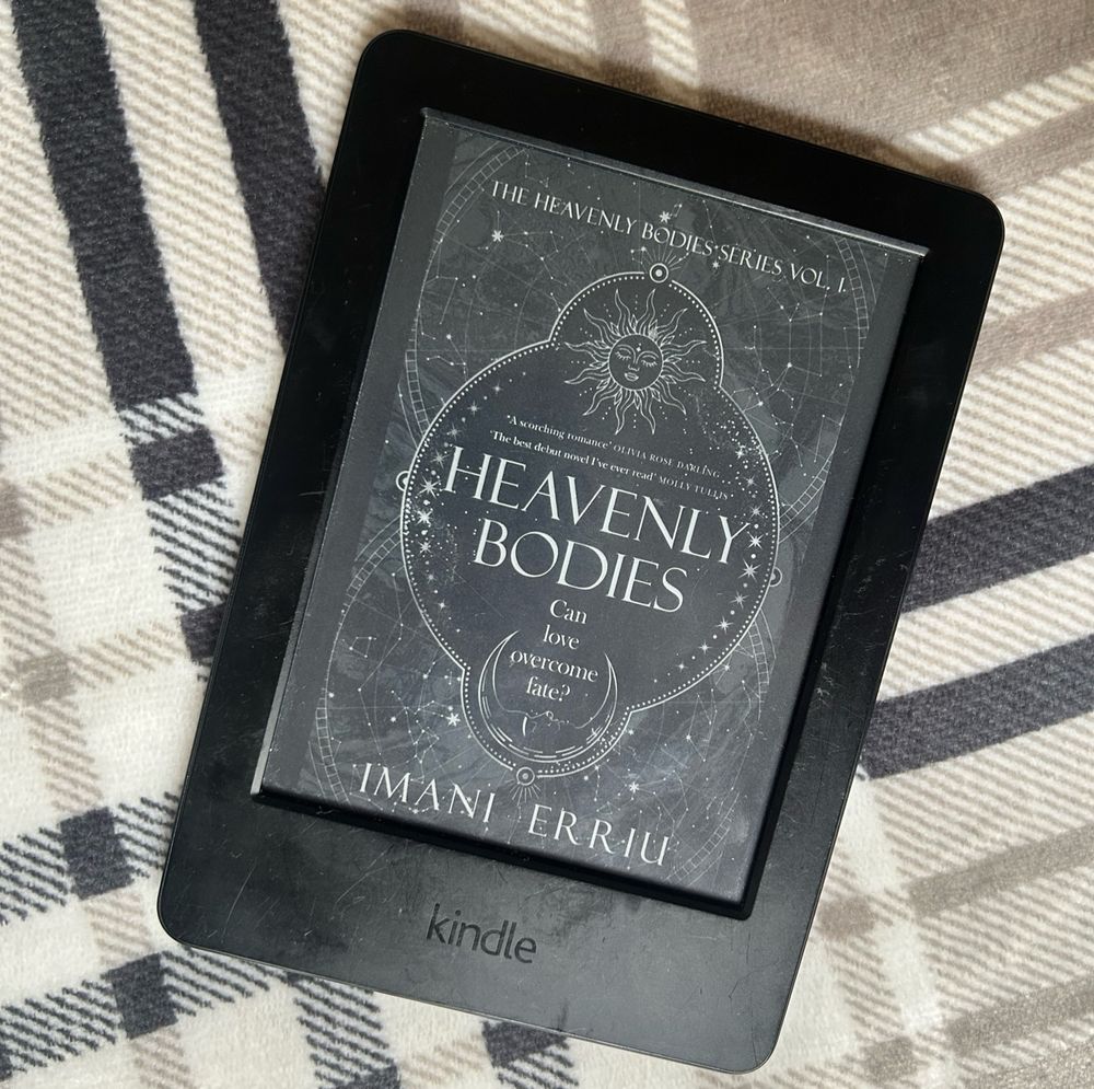 The digital cover of Heavenly Bodies, by Imani Erriu, on a kindle screen over a check background.