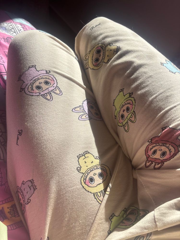 Recorte onde aparece somente as coxas vestindo calça de pijama. O pijama é de cor bege, com labubus desenhados espalhados em várias direções. Cada um tem uma cor diferente e pastel: amarelo, rosa, lilás, verde, marrom, cinza.