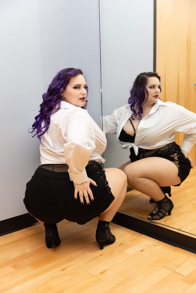 Photo of EnergeticJen squatting by a mirror with her hand on her butt and looking back over her shoulder wearing a sexy black lacy bodysuit, black shorts, black high heels, and a white button-up shirt tied in the front.