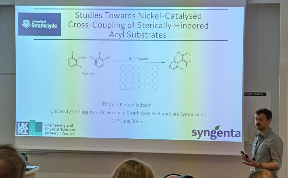 Image of a title slide for a presentation on "studies towards nickel-catalysed cross-coupling of sterically-hindered aryl substrates", with the presenter (Kieran Redpath) standing next to it.