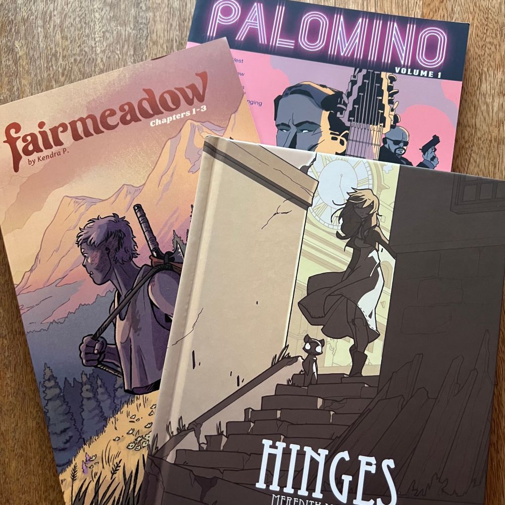 A collection of three books on a table: Palomino volume one by Stephan Franck, Fairmeadow chapters 1-3 by Kendra P., and Hinges book one by Meredith McClaren.