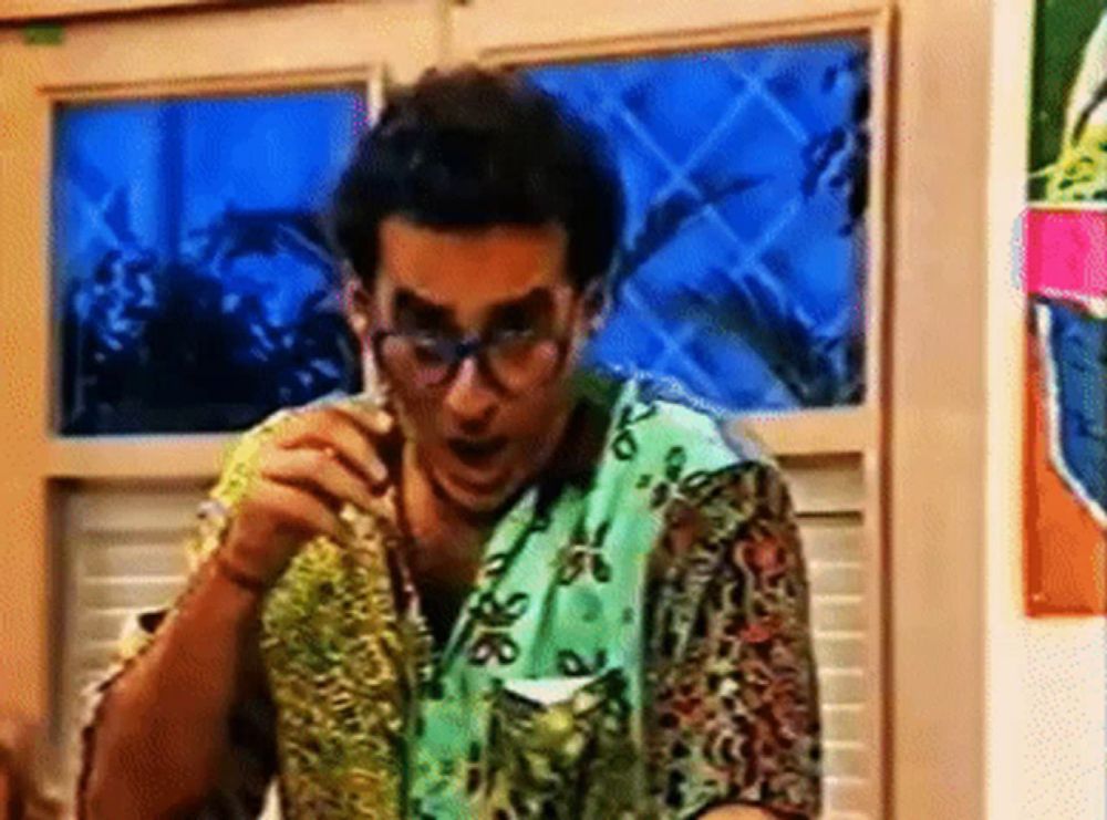 a man wearing glasses and a colorful shirt is sitting in front of a window eating a sandwich .