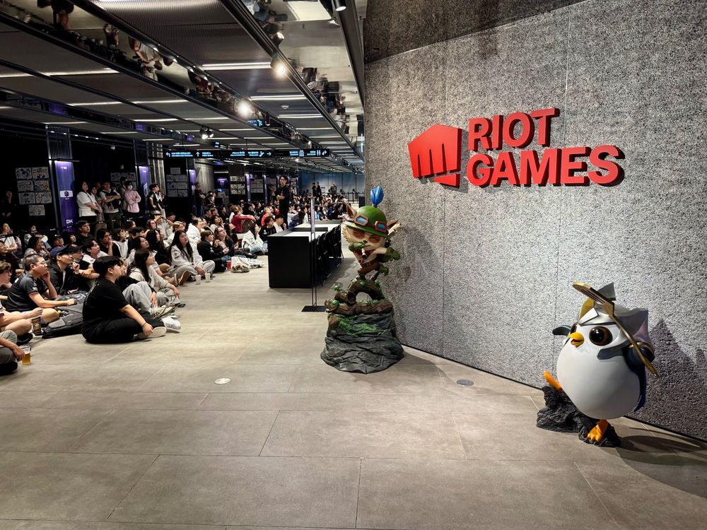 LoL Park during the series between T1 and Dplus Kia. On the right are statues of Teemo and Pengu, either side of a Riot Games sign. On the left, audience overflow for fans who cannot watch from the arena; fans are seated on the floor or standing in the back watching screens showing the game (screens not pictured)