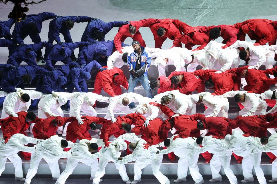Photo of Kendrick Lamar at the Super Bowl Halftime show, standing in the middle of an American flag made by Black people bending over, dressed in the colors of red, white, and blue, symbolizing that America was built off the backs of Black folks 👏🏾👏🏾👏🏾