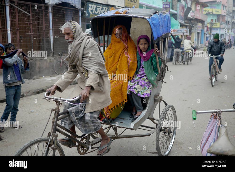 An old man is pedaling on a 3 wheeled extended bike with a cabin attached with a woman and girl in open cabin