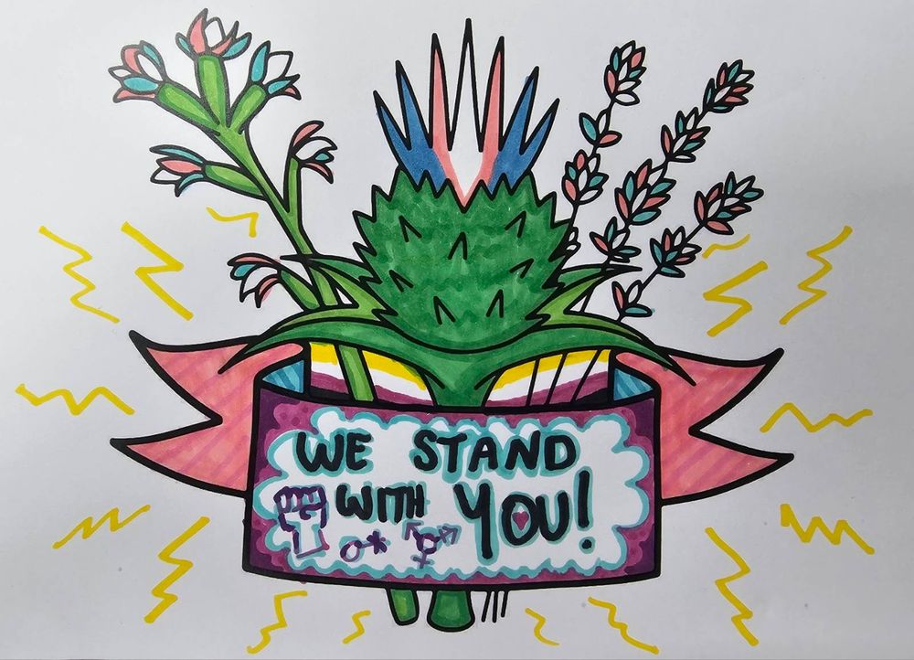 A coral root orchid and thistle have beautifully shaded green stalks and are topped with flowers in blue, pink and white with the trans pride colours continuing into the lavender flowers. The banner is pink and purple with blue accents and a message that reads "we stand with you!" There is a tiny heart hidden in the O of the you and trans symbols with a little fist drawn next to the message. A nonbinary flag is slipped in the space between the banner and the flowers and the whole design is surrounded by yellow zigzags that look like lightning bolts.