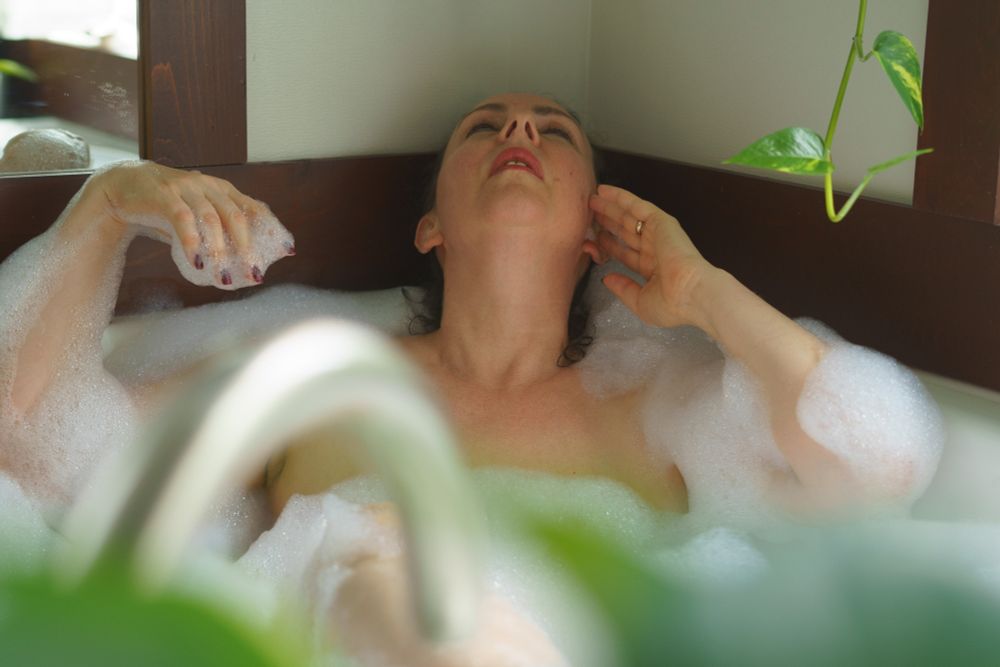 Athena lounges in a wide bathtub surrounded by foliage and woodgrains. Her head is laid back and her eyes are closed. Bubbles cling to her fingertips, arms and cover her torso and legs