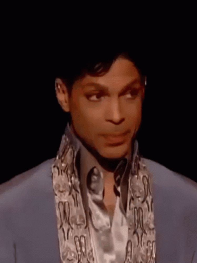 prince is wearing a blue suit and a scarf around his neck and making a funny face .