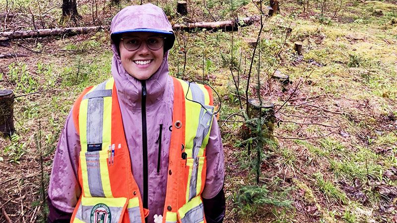 Smiling person wearing a reflective vest and a helmet standing in a wooded area.