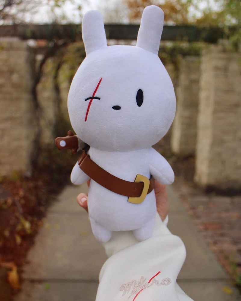 A photo of a plush of a white scarred bunny. The bunny is wearing a leather belt and a knife. The background of the photo has rustic-looking columns and autumn-colored trees. 