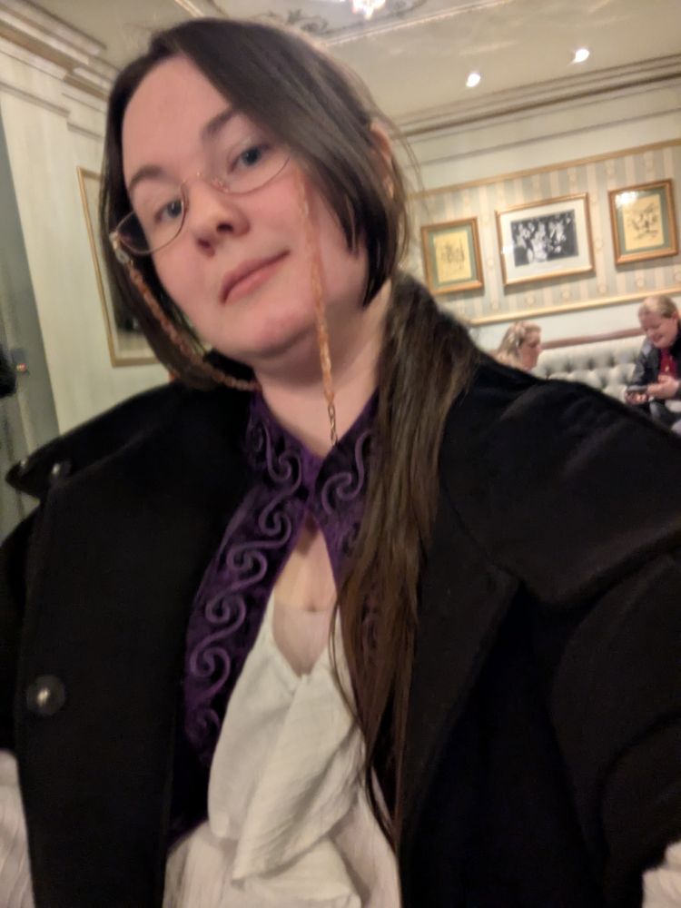 Photo of a brunette person wearing half-moon glasses and vaguely gothic clothing smiling lightly at the camera