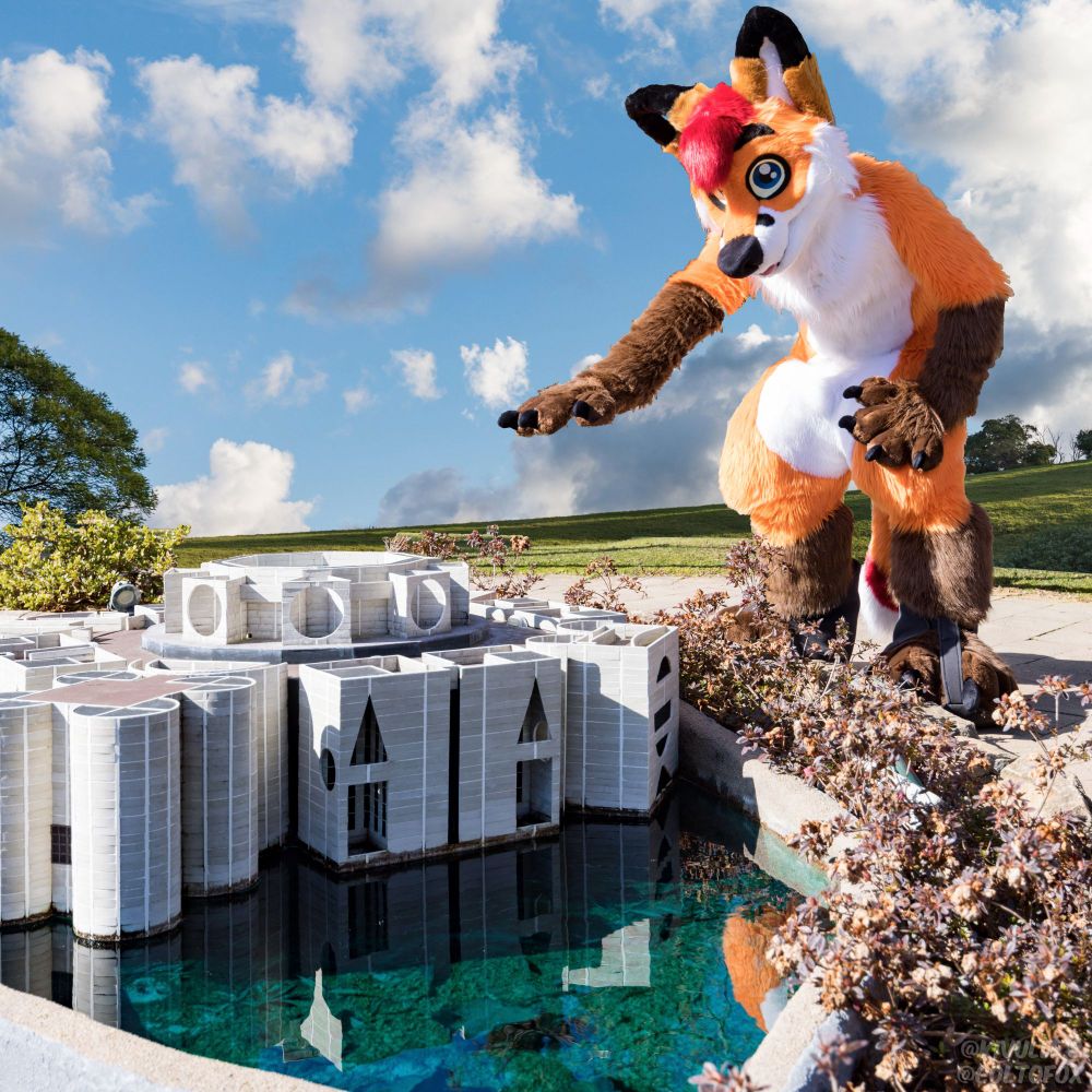 A photo of a castle surrounded by water and bushes on the shore. A gigantic Colto Fox (orange, white, brown and red fox fursuiter) is leaning over the castle with his right paw reaching down. There are clouds and blue sky in the background with a green grassy hill.