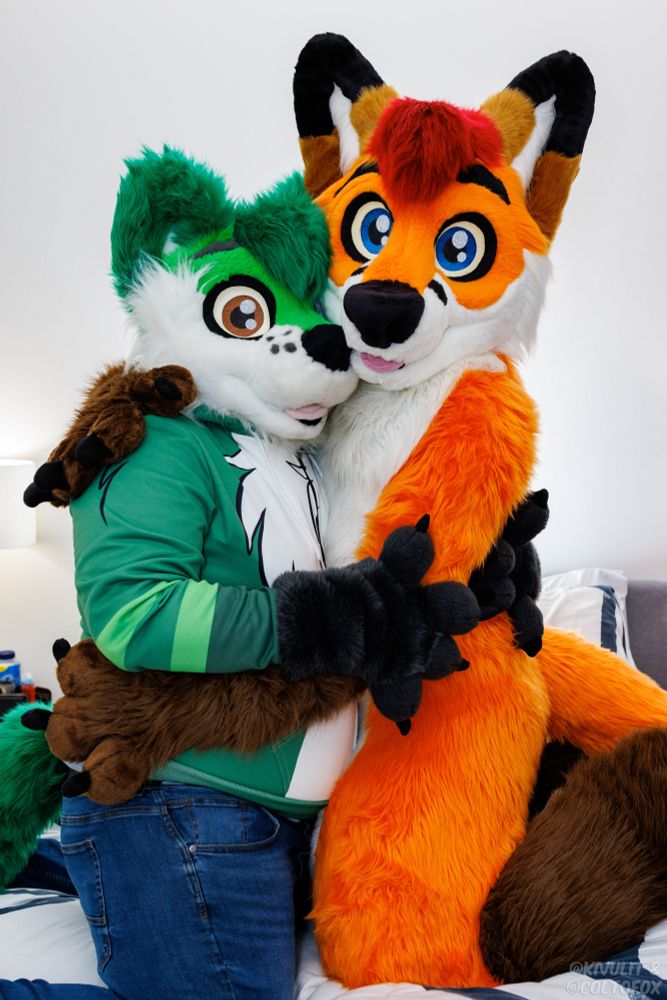 Makitoba (green and white otter-fox fursuiter) in partial fursuit kneeling on a bed wearing a green and white jacket hugging Colto Fox (orange, white, brown and red fox fursuiter) in full fursuit.