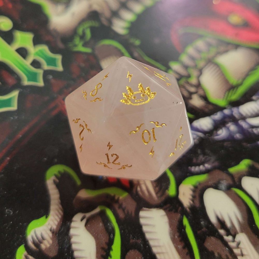 Close up of rose quartz d20 die with the face of an axolotl as the 20 in a gold paint inlay. It sits on a tray that has a skeleton with an open mouth and pierced tongue out.