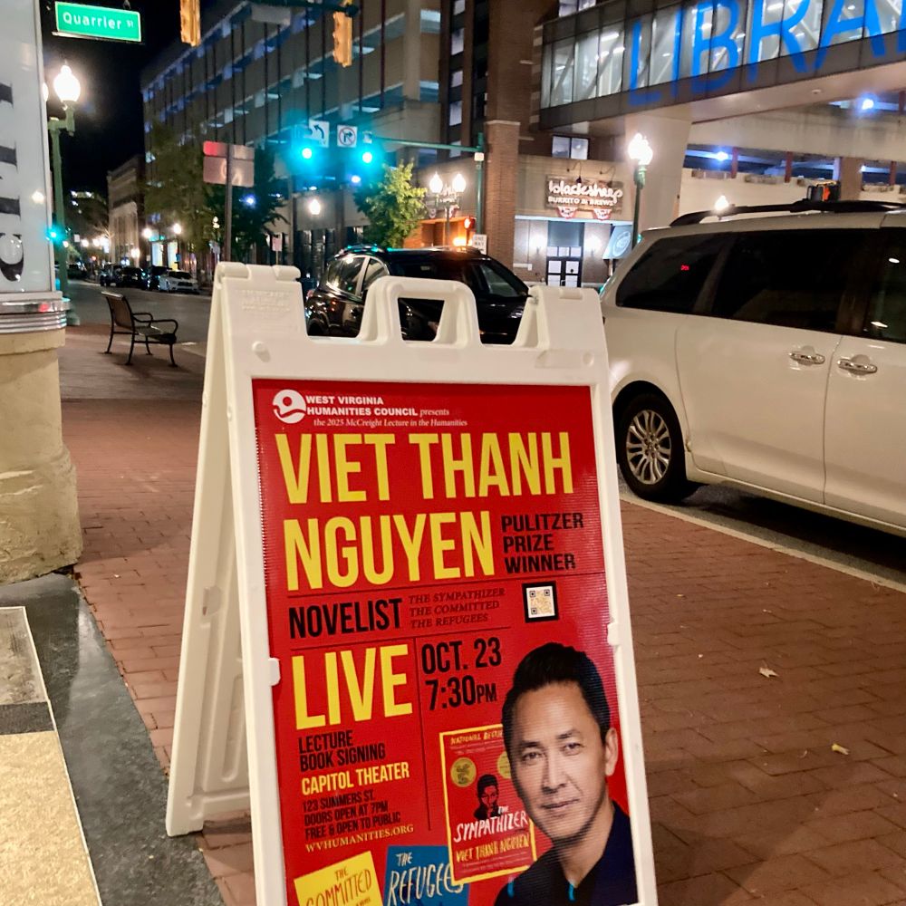 Sandwich board sign advertises appearance of Pulitzer Prize-winning novelist Viet Thanh Nguyen at 7:30 pm Oct. 23 and features his face, along with covers of three books: “The Sympathizer,” “The Refugees,” and “The Committed.” The sign is on a sidewalk near an intersection with a green traffic light at night in downtown Charleston, W.Va.