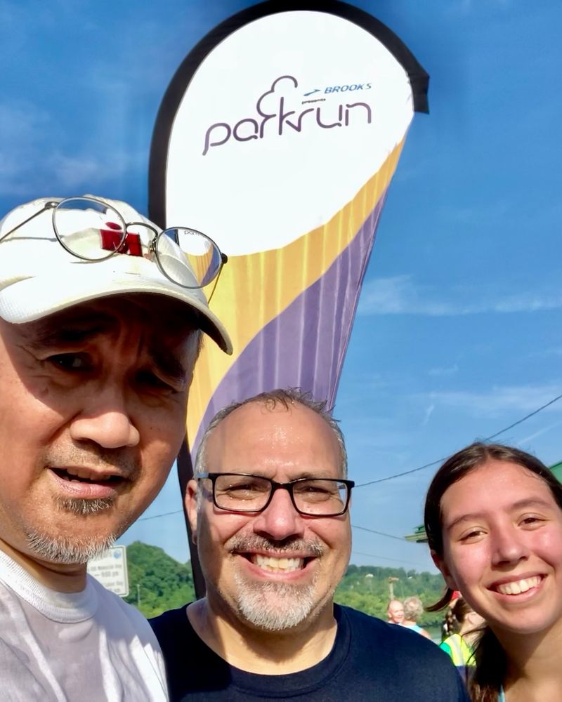 Selfie of middle-aged man with his friend and his daughter. A Parkrun banner is in the background. 