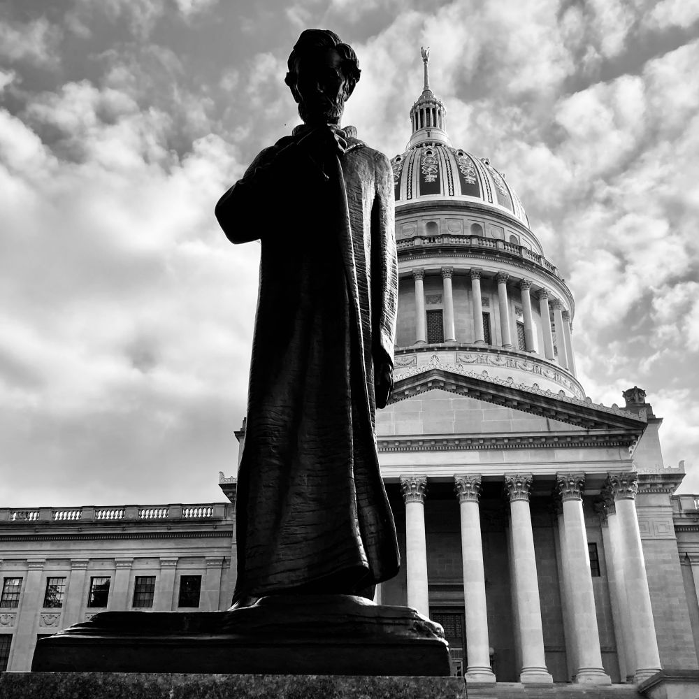 Statue of Abraham Lincoln wearing a long cloak. In the background is the dome of the West Virginia Capitol. 