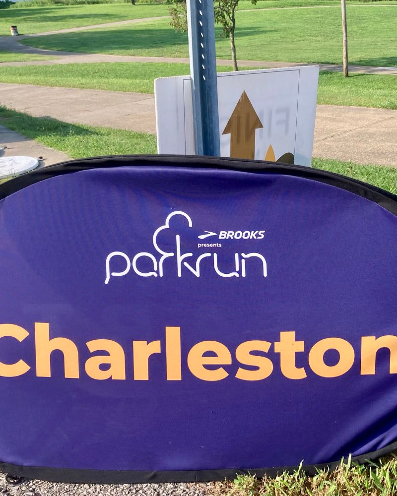 Charleston WV Parkrun popup sign with a Brooks sponsorship logo. A park and walking path is in the background. 