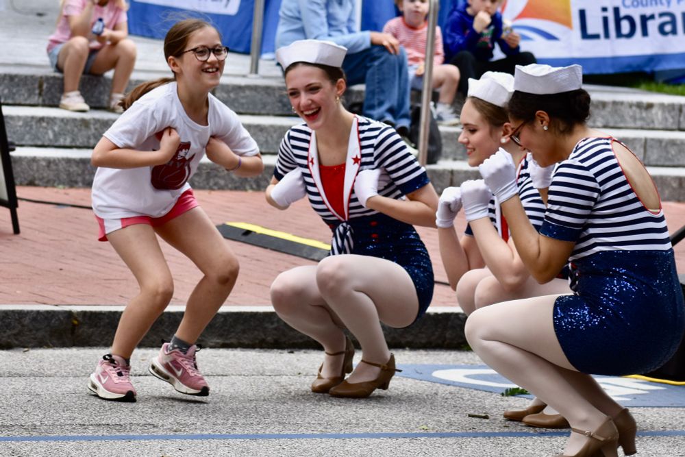 A little girl wearing shorts and a t-shirt tucks her arms like a duck and squats along with three teenage tap dancers wearing striped, sequined sailor suits. 