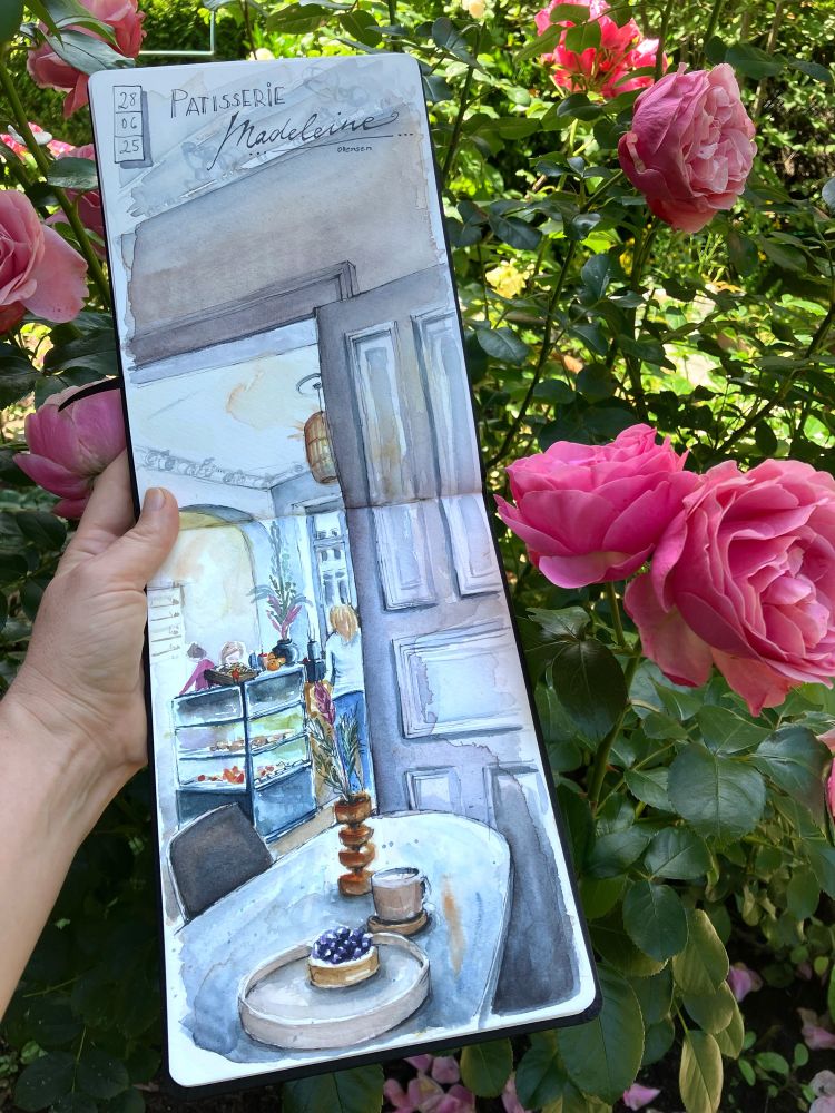 Open sketchbook showing sketch of the inside of a coffee place,blueberry cake in the foreground, counter and people in the background. 