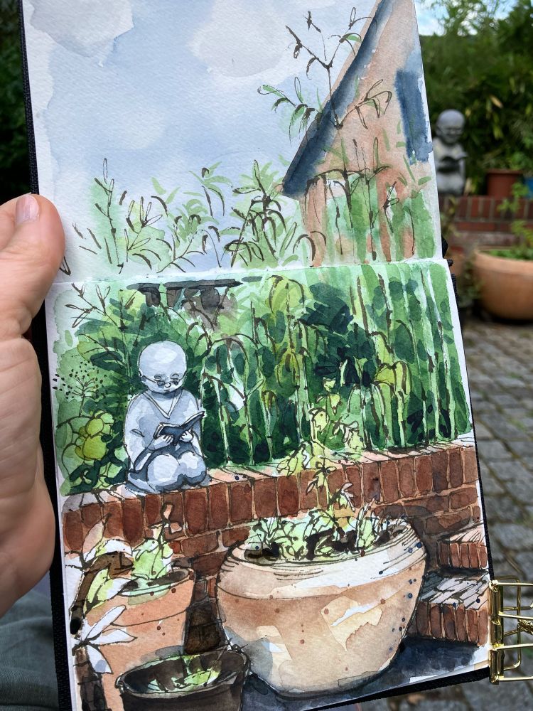 sketchbook showing sketch of garden plants, a little Buddha sculpture and a bamboo hedge 