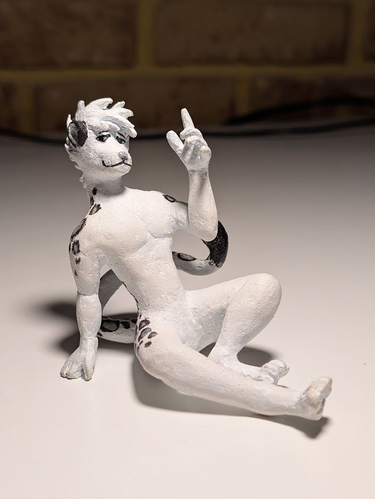 A handmade figurine of Zyyph - a snow leopard fursona.
The figure is sitting down and leaning back with one hand help up and other bracing against the ground. It is facing towards the viewer. A painted and finished figurine.